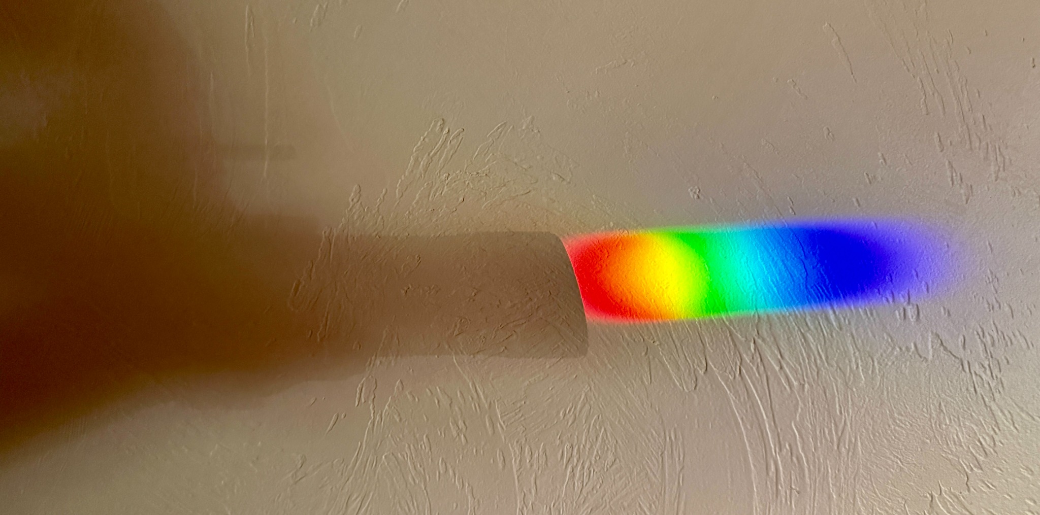 A band of vivid prismatic light emerging from shadow across a textured wall